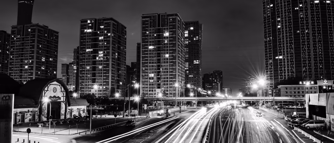 Long-exposure traffic light trails streak through a dense city skyline lit up after dark.